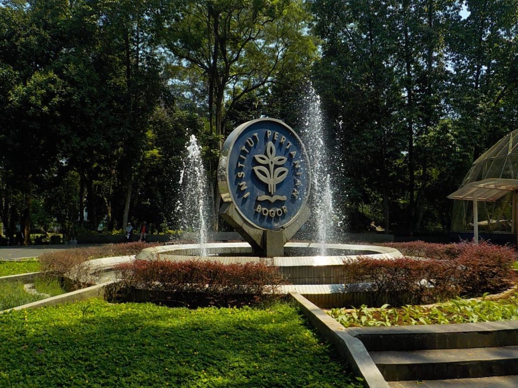 Fountain with the Institut Pertanian Bogor emblem surrounded by vibrant greenery and trees.