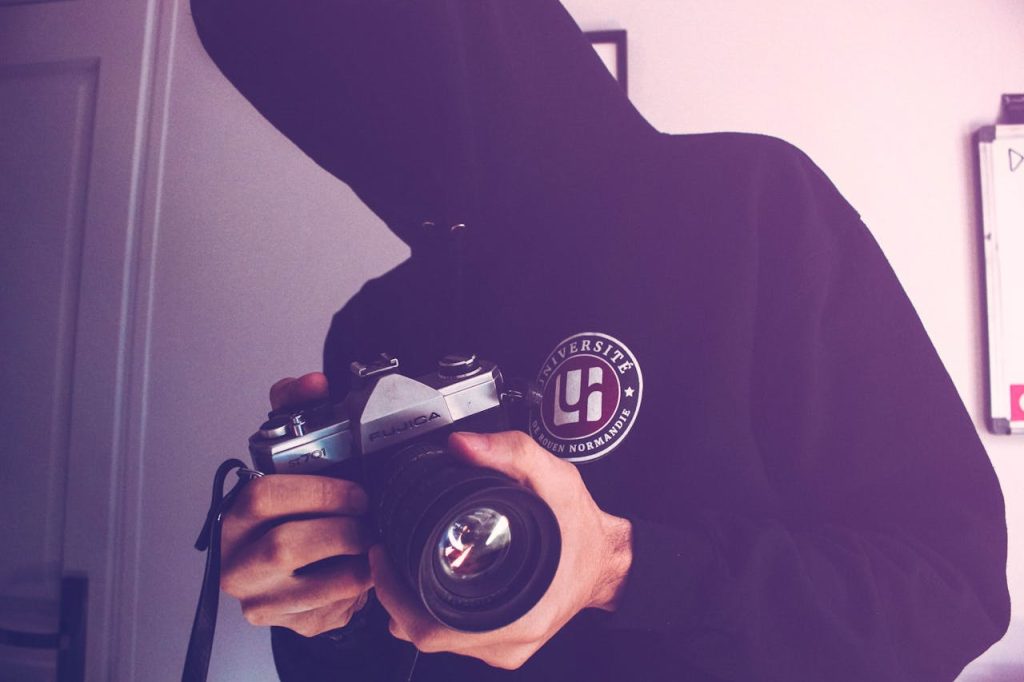 A hooded individual holding a vintage camera indoors, capturing a classic photography moment.