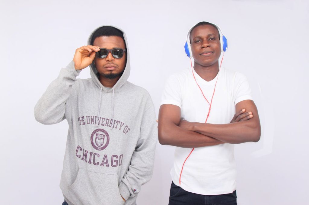 Two young men in casual attire, one wearing sunglasses and a hoodie, the other with headphones, against a white background.